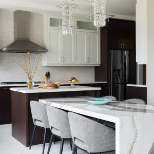 A bright, luxurious modern kitchen featuring a multi-level island design. The lower tier consists of a stunning integrated dining table with a thick white and grey marble waterfall edge, accompanied by grey upholstered armchairs with black legs. Above the table hang two contemporary geometric chrome and crystal cylinder pendant lights. The higher tier island features a dark wood base with a white quartz counter. The background shows two-tone cabinetry, a full-height stone backsplash, a stainless steel range hood, and a black French door refrigerator.