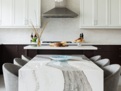 A spacious modern kitchen showcasing a unique two-tier island setup. In the foreground, a low-profile dining table extension features a dramatic white quartz or marble waterfall edge with bold, wide grey veining, surrounded by plush grey upholstered dining chairs. Behind it, the elevated main kitchen island has a crisp white countertop styled with fresh bread and pastries. The background reveals two-tone cabinetry with dark espresso base units and white shaker upper cabinets, a full-height stone backsplash, and a sleek stainless steel range hood.