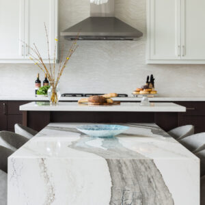 A spacious modern kitchen showcasing a unique two-tier island setup. In the foreground, a low-profile dining table extension features a dramatic white quartz or marble waterfall edge with bold, wide grey veining, surrounded by plush grey upholstered dining chairs. Behind it, the elevated main kitchen island has a crisp white countertop styled with fresh bread and pastries. The background reveals two-tone cabinetry with dark espresso base units and white shaker upper cabinets, a full-height stone backsplash, and a sleek stainless steel range hood.