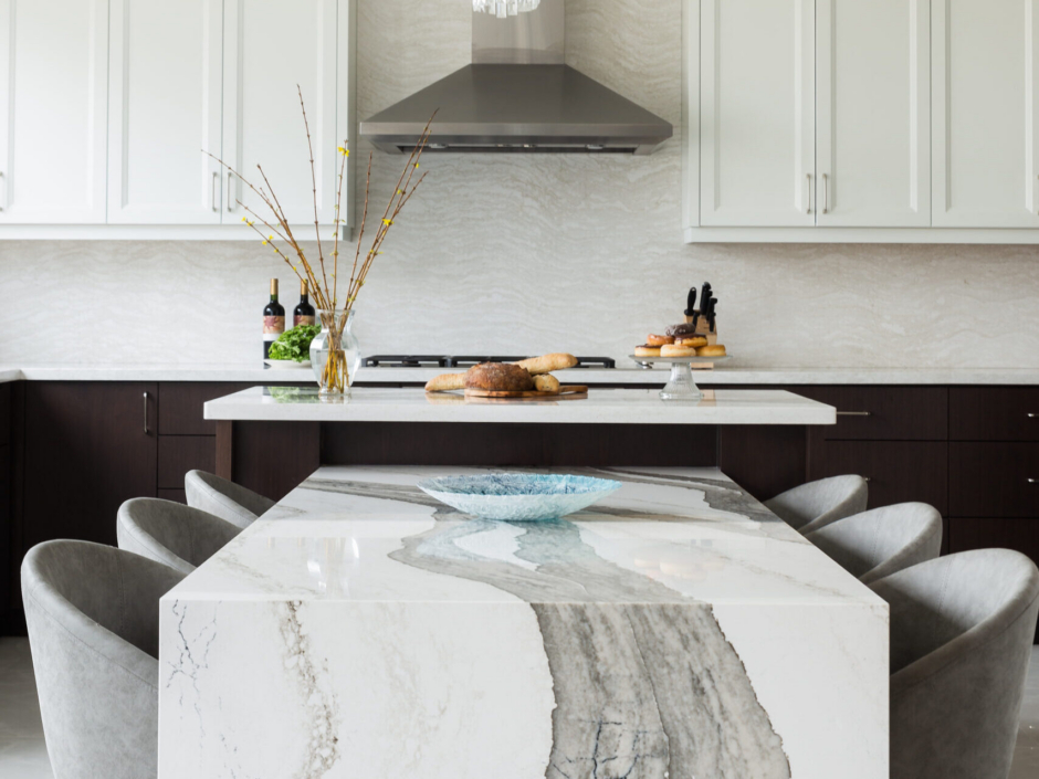A spacious modern kitchen showcasing a unique two-tier island setup. In the foreground, a low-profile dining table extension features a dramatic white quartz or marble waterfall edge with bold, wide grey veining, surrounded by plush grey upholstered dining chairs. Behind it, the elevated main kitchen island has a crisp white countertop styled with fresh bread and pastries. The background reveals two-tone cabinetry with dark espresso base units and white shaker upper cabinets, a full-height stone backsplash, and a sleek stainless steel range hood.