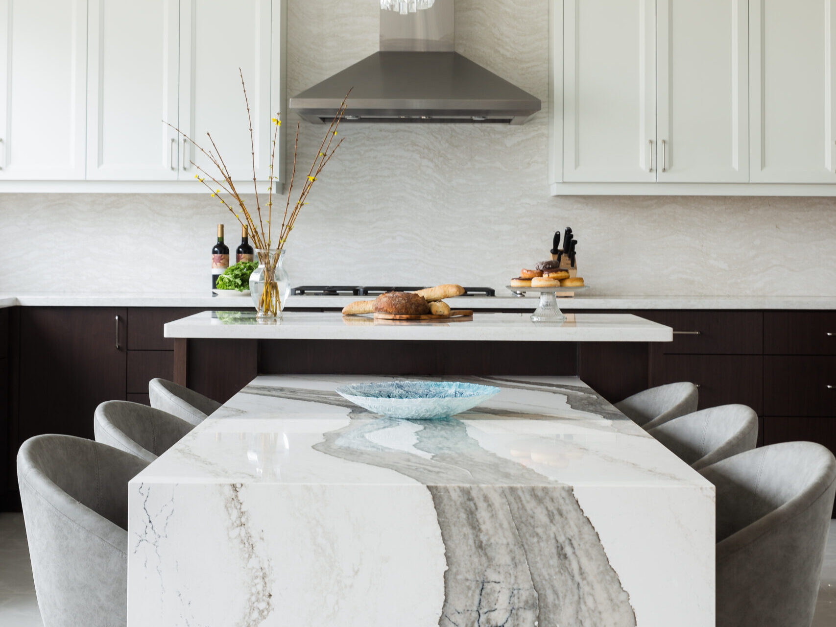 A spacious modern kitchen showcasing a unique two-tier island setup. In the foreground, a low-profile dining table extension features a dramatic white quartz or marble waterfall edge with bold, wide grey veining, surrounded by plush grey upholstered dining chairs. Behind it, the elevated main kitchen island has a crisp white countertop styled with fresh bread and pastries. The background reveals two-tone cabinetry with dark espresso base units and white shaker upper cabinets, a full-height stone backsplash, and a sleek stainless steel range hood.