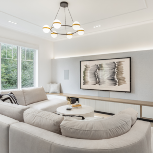 A spacious modern living room featuring a large beige curved sectional sofa facing a custom, wall-to-wall built-in media console. The sleek console combines a light wood top with white flat-panel and fluted cabinet doors, extending into a cozy window bench. A large flat-screen TV displaying abstract art is mounted on a lightly textured, backlit accent wall, flanked by flush in-wall speakers. A striking modern black and brass ring chandelier with white glass globes hangs from the ceiling, illuminating the bright space.