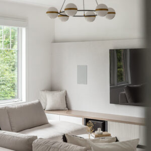 A bright modern living room featuring a cozy light grey sectional sofa facing a custom built-in media console. The console showcases a light wood top over white drawers and textured fluted paneling, extending into a window bench with a patterned throw pillow. A large flat-screen TV and a sleek grey in-wall speaker are mounted on a lightly textured accent wall. Above, a striking modern chandelier with a black ring, brass accents, and white glass globes illuminates the space.