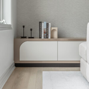 A close-up view of a modern living room featuring a custom low-profile built-in console. The sleek cabinet combines a light taupe frame with a distinct curved corner and flat white doors, resting on a black recessed base. The surface is minimally styled with slender black candlesticks, a stack of art books, and a wooden arch object, all set against a textured light grey wall just behind a white sofa.