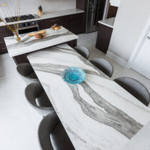 A high-angle, overhead view of a luxury modern kitchen highlighting an integrated multi-level island. The prominent lower dining table extension showcases a dramatic white slab surface with thick, sweeping charcoal grey veins, styled with a turquoise glass bowl and surrounded by six grey upholstered tub chairs. The upper-tier kitchen island features a matching veined countertop with bakery items. The surrounding kitchen area is designed with dark wood perimeter base cabinets, light grey upper cabinets, and a stainless steel French door refrigerator.