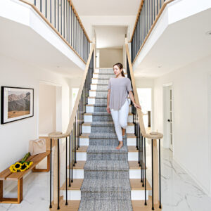 A woman walking barefoot down a grand, straight staircase in a bright, modern two-story foyer. The stairs feature light wood treads, white risers, and a textured grey patterned carpet runner. The railing consists of a light wood handrail with curved volute ends and sleek black metal balusters that continue along the upper hallway balcony. The entryway is finished with polished white marble-look floor tiles and white walls. To the left, a wooden bench holds a canvas tote bag and fresh sunflowers beneath a framed landscape photograph.