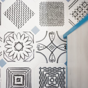 A top-down, close-up view of a bathroom floor featuring patterned white tiles adorned with various black hand-drawn geometric, floral, and line-art motifs. The main patterned square tiles are bordered by small, light blue accent corner tiles. To the right, a blurred edge of a floating light wood vanity with a vibrant cobalt blue top trim frames the shot.