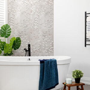 A close-up view of a bright, modern bathroom featuring a sleek white freestanding bathtub with a textured dark blue towel draped over its edge. A striking grey and white chevron mosaic tile accent wall serves as a backdrop, complemented by a contemporary matte black floor-mounted tub filler. The spa-like space is accented with a large faux monstera plant to the left, while the crisp white wall to the right shows a partial view of a black heated towel rack.