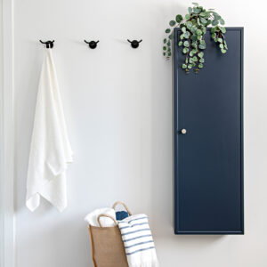 A bright, minimalist bathroom wall featuring three matte black towel hooks in a row, with a fluffy white towel hanging on the leftmost hook. To the right is a tall, flat-panel navy blue storage cabinet accented with a single silver knob and topped with a trailing green plant. On the floor below the hooks, a woven basket holds a neat arrangement of rolled white and blue striped towels.