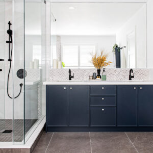 A bright, modern bathroom featuring a long navy blue double vanity with silver knob hardware and a crisp white quartz countertop. Above the under-mount sinks, a grey and white chevron mosaic tile backsplash runs beneath a large, frameless rectangular mirror. Matte black faucets match the fixtures visible in the adjacent frameless glass walk-in shower on the left, which has a dark grey tiled floor. The vanity is styled with a vase of dried yellow florals and various bathroom accessories.