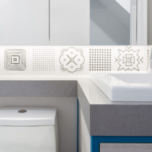 A close-up view of a modern bathroom highlighting a decorative backsplash made of white square tiles with unique black and grey geometric patterns, illuminated by under-cabinet LED lighting. The tile band runs above a thick grey quartz countertop that wraps into a corner over a white toilet. On the right, a white drop-in sink with a polished chrome faucet sits on the vanity counter, accented with a tiny potted succulent.