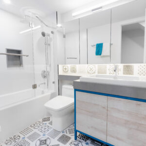 A bright, modern bathroom featuring an eye-catching blue and white patterned tile floor with geometric motifs. In the foreground, a light wood floating vanity is outlined with a striking cobalt blue frame and topped with a thick grey stone counter and white vessel sink. A large multi-panel mirror with integrated lighting sits above an intricate patterned tile backsplash. To the left, a clean white alcove bathtub is enclosed by a frameless glass shower panel and equipped with polished chrome fixtures.