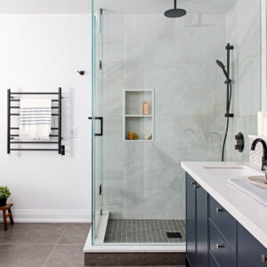 A modern bathroom featuring a frameless glass walk-in shower with large-format light grey stone-look wall tiles, a dark mosaic tile floor, and matte black shower fixtures. The shower includes a white built-in double niche holding bath products. To the left, a matte black heated towel rack holds a white and blue striped towel against a crisp white wall. To the right, a partial view of a navy blue vanity is visible, complete with a white countertop, under-mount sink, and a matte black faucet.
