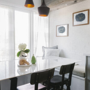 A bright, modern breakfast nook featuring a white marble dining table, two industrial-style black metal chairs, and a built-in white banquette bench with cozy pillows. The space is set against a textured white painted brick wall adorned with framed constellation art, sitting below a rustic reclaimed wood ceiling beam. A cluster of three black pendant lights with warm gold interiors hangs above the table, which is styled with fresh white hydrangeas, a glass cloche, and pastries near a large window with sheer white curtains.