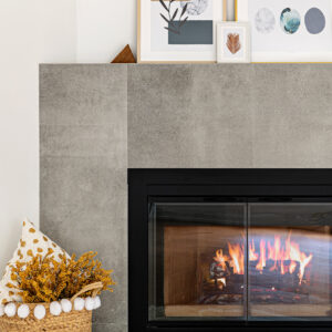 A close-up view of a modern fireplace featuring a sleek grey concrete-look tile surround and a matte black metal frame with a protective mesh screen. A warm wood fire burns brightly inside. The hearth area to the left is styled with a white and gold polka dot throw pillow alongside a basket of dried yellow florals, while framed artwork leans casually on the mantle above.