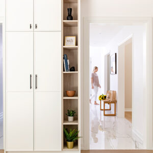 A bright, modern hallway featuring custom floor-to-ceiling white flat-panel storage cabinets with matte black pulls, adjacent to a tall, light wood open shelving unit styled with books, decor, and small plants. Through an open doorway to the right, a woman walks barefoot down a sunlit corridor with polished white marble-look floors, passing a small wood and metal bench holding a shopping bag and a bouquet of fresh yellow sunflowers.