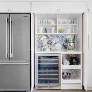 A modern kitchen wall featuring a stainless steel French door refrigerator alongside tall white built-in cabinetry. The cabinet doors are open to reveal a hidden beverage center and appliance garage. The illuminated interior features open shelving styled with dishware, a counter workspace with a mint green stand mixer, and a vibrant blue and white hexagon tile backsplash. Below the counter sits an integrated stainless steel wine fridge next to convenient pull-out pantry drawers.