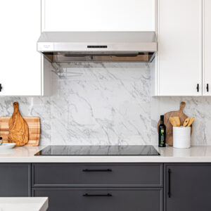 A close-up view of a modern two-tone kitchen featuring a sleek black induction cooktop seamlessly integrated into a crisp white quartz countertop. Below the counter are dark grey flat-panel base cabinets with matte black pulls, contrasting with the bright white upper cabinets. A prominent stainless steel range hood sits above a striking, full-height white marble-look slab backsplash with grey veining. The space is warmly styled with leaning wooden cutting boards, a bottle of olive oil, and a textured white ceramic crock holding wooden cooking utensils.