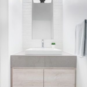 A bright, modern powder room featuring a floating light wood vanity with a thick grey stone countertop and a crisp white square vessel sink. A tall, frameless rectangular mirror is centered on a white painted brick accent wall and illuminated by a contemporary black and chrome sconce. The minimalist space is completed with a sleek chrome single-hole faucet, a small green potted plant, and a light grey towel hanging on the adjacent white wall.