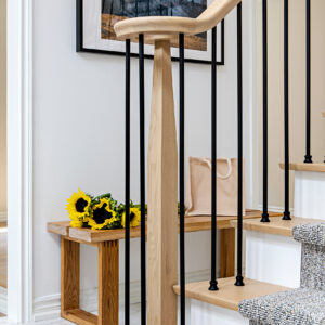 A close-up view of a modern staircase railing featuring a tapered light wood newel post and sleek matte black metal balusters. Through the spindles, a rustic light wood entryway bench is visible against a bright white wall, styled with a canvas tote bag and a fresh bouquet of bright yellow sunflowers. The staircase itself highlights light wood treads, crisp white risers, and a textured grey patterned carpet runner.