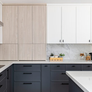 A bright, modern two-tone kitchen featuring crisp white upper cabinets and dark grey base cabinets, both accented with minimalist matte black hardware. A seamless white marble slab with grey veining forms the countertop and full-height backsplash under a stainless steel range hood. In the foreground, a dark grey kitchen island with a white countertop features a matte black gooseneck faucet, an under-mount sink, and light grey upholstered bar stools with light wood legs. Built-in stainless steel double wall ovens are visible to the left, and the space is styled with fresh fruit and greenery.