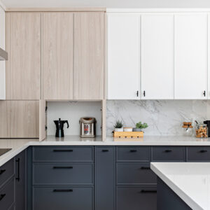 A modern three-tone kitchen design featuring dark grey base cabinets, crisp white upper cabinets, and tall, flat-panel light wood cabinetry. The lower door of the wood cabinetry is open to reveal a convenient hidden appliance garage resting directly on the white quartz countertop, housing a black moka pot and a stainless steel water boiler. To the right, a seamless white marble-look slab backsplash sits behind the styled counter area, which includes a wooden tray of small potted plants and glass storage jars. Minimalist matte black hardware ties the two-tone cabinetry together.