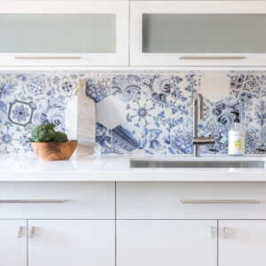 A close-up view of a modern two-tone kitchen featuring a vibrant blue and white patterned hexagon tile backsplash. Crisp white upper cabinets with horizontal frosted glass inserts sit above a seamless white solid-surface countertop. Below, sleek light grey flat-panel base cabinets are finished with brushed nickel pulls. The counter is styled with an under-mount stainless steel sink, a modern gooseneck faucet, a wooden bowl holding fresh broccoli, and a marble cutting board leaning against the colorful tiles.