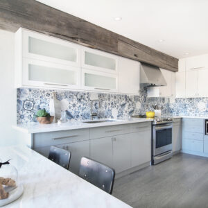 A bright, transitional two-tone kitchen featuring light grey flat-panel base cabinets and white upper cabinets, including a row of horizontal frosted glass doors. A striking blue and white patterned hexagonal tile backsplash runs along the wall beneath a prominent rustic reclaimed wood ceiling beam. The kitchen is equipped with sleek white countertops, a stainless steel range, and a matching hood vent. In the foreground, a marble-look peninsula with two black metal barstools is styled with a glass cloche and fresh fruit, set against grey wood flooring.
