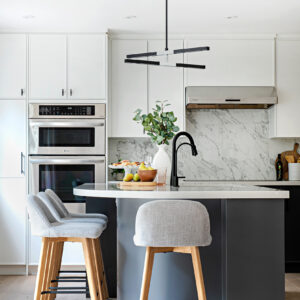 A bright, modern two-tone kitchen featuring crisp white flat-panel cabinetry and a contrasting dark grey island. A seamless white marble-look quartz forms the perimeter countertop and full-height backsplash beneath a stainless steel range hood. Built-in stainless steel double wall ovens are seamlessly integrated into the tall cabinets on the left. The curved kitchen island features a matte black gooseneck faucet, light grey upholstered bar stools with wood legs, and is illuminated from above by a contemporary black linear LED chandelier. The space is styled with fresh fruit, pastries, and a white vase holding greenery.