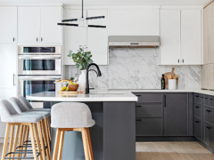 A bright, modern two-tone kitchen featuring crisp white upper cabinets and dark grey base cabinets, both accented with minimalist matte black hardware. A seamless white marble slab with grey veining forms the countertop and full-height backsplash under a stainless steel range hood. In the foreground, a dark grey kitchen island with a white countertop features a matte black gooseneck faucet, an under-mount sink, and light grey upholstered bar stools with light wood legs. Built-in stainless steel double wall ovens are visible to the left, and the space is styled with fresh fruit and greenery.