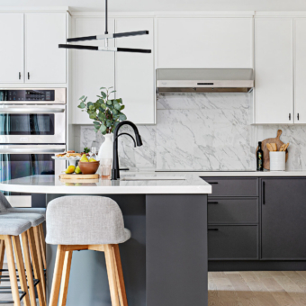 A bright, modern two-tone kitchen featuring crisp white upper cabinets and dark grey base cabinets, both accented with minimalist matte black hardware. A seamless white marble slab with grey veining forms the countertop and full-height backsplash under a stainless steel range hood. In the foreground, a dark grey kitchen island with a white countertop features a matte black gooseneck faucet, an under-mount sink, and light grey upholstered bar stools with light wood legs. Built-in stainless steel double wall ovens are visible to the left, and the space is styled with fresh fruit and greenery.