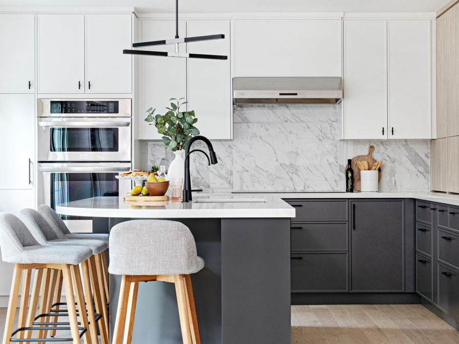 A bright, modern two-tone kitchen featuring crisp white upper cabinets and dark grey base cabinets, both accented with minimalist matte black hardware. A seamless white marble slab with grey veining forms the countertop and full-height backsplash under a stainless steel range hood. In the foreground, a dark grey kitchen island with a white countertop features a matte black gooseneck faucet, an under-mount sink, and light grey upholstered bar stools with light wood legs. Built-in stainless steel double wall ovens are visible to the left, and the space is styled with fresh fruit and greenery.