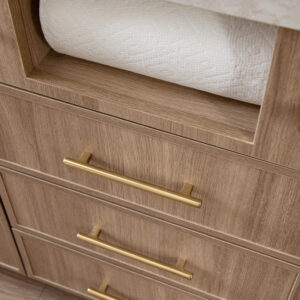 A close-up view of custom light wood kitchen cabinetry featuring a built-in open slot holding a roll of white paper towels. Below the paper towel holder are three drawers equipped with brushed gold bar pulls, all situated underneath a light patterned quartz countertop.