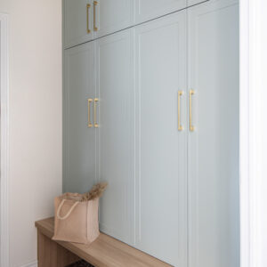 A modern mudroom entryway featuring floor-to-ceiling dusty blue built-in storage cabinets adorned with brushed brass bar hardware. Below the cabinetry is a custom light oak wood bench serving as seating and open storage. The space is grounded by decorative geometric patterned floor tiles, with a canvas tote bag resting on the bench and a pair of grey shoes tucked underneath.