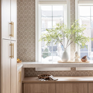 A bright, modern kitchen interior featuring light oak built-in cabinets with brushed gold hardware. The wall is accented with a taupe scallop-patterned tile backsplash. A white quartz countertop extends across large windows, displaying a white vase with leafy branches, while a built-in wooden storage bench sits below.