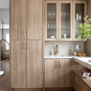 A modern built-in wet bar and pantry area featuring light oak wood cabinetry with brushed gold hardware. The setup includes a tall storage cabinet, lower base cabinets, and upper cabinets with fluted glass doors displaying glassware. A small prep sink with a gold faucet sits nestled in a white quartz countertop, complementing the warm light wood flooring throughout the space.
