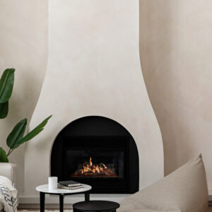 A cozy, minimalist living room corner featuring a tall, custom curved plaster fireplace surround with an arched black firebox and a lit fire. In front of the fireplace, resting on a fluffy white area rug, are two small round nesting tables and a modern taupe bean bag floor chair. A large green leafy houseplant and the edge of a white sofa frame the left side of the image against textured cream-colored walls.