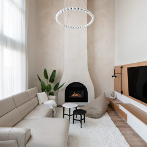 A wide view of a spacious, modern living room with a double-height ceiling featuring a warm wood slat detail. A tall, custom curved white plaster fireplace serves as the focal point, with a striking circular globe chandelier hanging above. The cozy, neutral-toned space is furnished with a cream leather sectional sofa, a fluffy white shag area rug, a taupe bean bag chair, and nesting coffee tables. To the right is a custom wood-paneled media wall with a floating console, and to the left, a large potted plant sits beside tall windows draped with sheer curtains.
