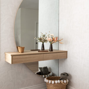 A modern entryway corner featuring a light wood floating console table with drawers, mounted across a large, frameless half-arch mirror. The space is styled with a subtle light grey abstract patterned wallpaper. On the console sit two small dark glass vases with dried florals, while a woven basket with white pom-poms rests on the light oak hardwood floor below.