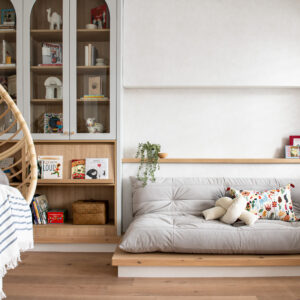 A cozy, modern children's reading nook featuring a hanging rattan egg chair with a striped throw blanket, and a low-profile grey floor cushion resting on a raised wood platform. The space includes custom built-in cabinetry with grey arched glass doors and open wood shelving filled with books. A shallow wood picture ledge displaying books and a trailing plant runs along the bright white wall above the seating area.