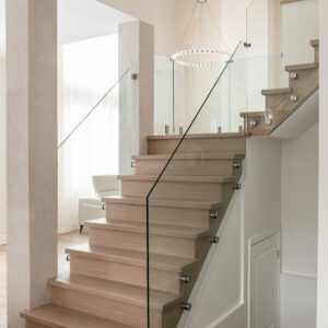 A modern light wood staircase featuring frameless glass railings secured with stainless steel standoff pins. The contemporary interior showcases textured beige pillars, a double-height ceiling with a striking circular globe chandelier, and a decorative wood slat ceiling detail in a bright, open-concept home.
