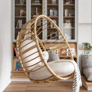 A detailed close-up of a modern children's playroom reading nook, featuring a hanging rattan egg chair with white cushions and a blue and white striped throw. The background shows custom light-grey arched glass-front built-in cabinets filled with kids' books and decor, above open shelves, on light oak flooring.