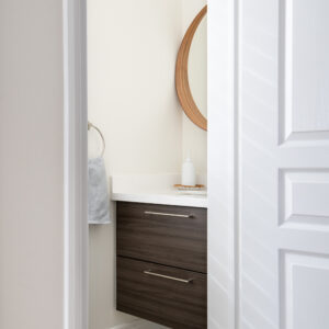 A peek through a partially open white paneled door into a bright, modern powder room. The small bathroom features a dark wood two-drawer floating vanity with a white countertop and a round wooden mirror. The floor is accented with blue and cream leaf-patterned hexagonal tiles, and a textured grey hand towel hangs on a silver ring against the cream-colored wall.