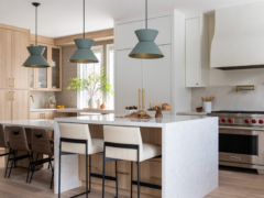 A wide view of a bright, modern two-tone kitchen featuring a large two-tier island with a white quartz waterfall countertop and light oak wood base. Three sage green geometric pendant lights hang above the island. The space blends crisp white and light wood cabinetry with brushed brass hardware, and includes a stainless steel gas range beneath a custom white plaster hood. White and brown leather counter stools provide seating around the island.