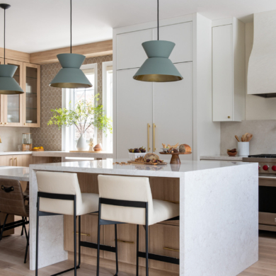 A wide view of a bright, modern two-tone kitchen featuring a large two-tier island with a white quartz waterfall countertop and light oak wood base. Three sage green geometric pendant lights hang above the island. The space blends crisp white and light wood cabinetry with brushed brass hardware, and includes a stainless steel gas range beneath a custom white plaster hood. White and brown leather counter stools provide seating around the island.