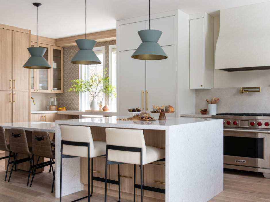 A wide view of a bright, modern two-tone kitchen featuring a large two-tier island with a white quartz waterfall countertop and light oak wood base. Three sage green geometric pendant lights hang above the island. The space blends crisp white and light wood cabinetry with brushed brass hardware, and includes a stainless steel gas range beneath a custom white plaster hood. White and brown leather counter stools provide seating around the island.