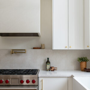A close-up of a warm, minimalist modern kitchen featuring a stainless steel gas range with red knobs situated beneath a custom, textured plaster range hood. A brass pot filler is mounted on a seamless, beige stone backsplash with a narrow integrated display ledge. To the right, crisp white cabinetry with brushed gold hardware surrounds a white quartz countertop and an apron-front farmhouse sink.
