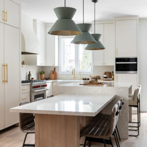 A bright, modern kitchen featuring a custom two-tier island with a high quartz waterfall counter and a lower light wood dining table extension. Three large sage green cone-shaped pendant lights hang above. The space includes cream-colored cabinetry with brass hardware, brown ribbed leather dining chairs, a custom white range hood, and light oak hardwood floors.