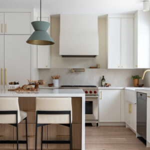 A bright, modern kitchen featuring a light wood island with a white quartz waterfall countertop and two white leather counter stools with black frames. A distinctive sage green geometric pendant light hangs above the island. The background showcases crisp white cabinetry with brass hardware, a custom white plaster range hood over a stainless steel gas stove, a brass pot filler, and light oak hardwood floors.