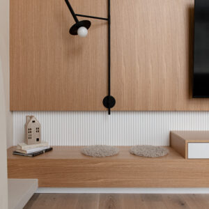 A close-up of a modern living room media wall featuring a large, light oak wood panel adorned with a matte black articulated wall sconce. Below the wood panel is a white fluted wall texture and a custom floating light wood bench. The bench is styled with two round textured seat cushions, a stack of books, and a small ceramic house decor piece. The edge of a black flat-screen TV is visible on the right, situated above light hardwood floors and a fluffy white area rug.